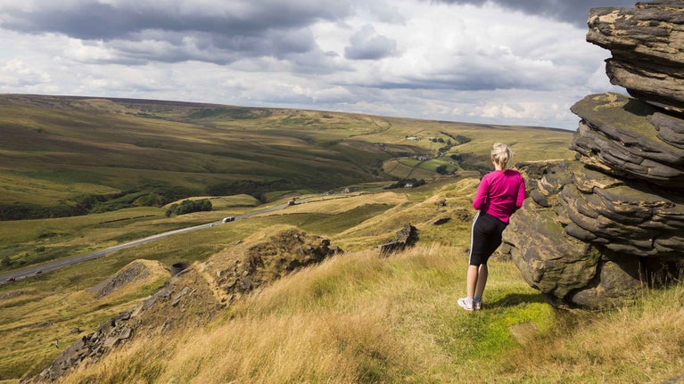 A woman is leaning against a huge craggy rock with her back to the camera, admiring the view from Pule Hill on Marsden Moor, West Yorkshire. Grey clouds cast shadows across the moor, which stretches for miles into the distance.